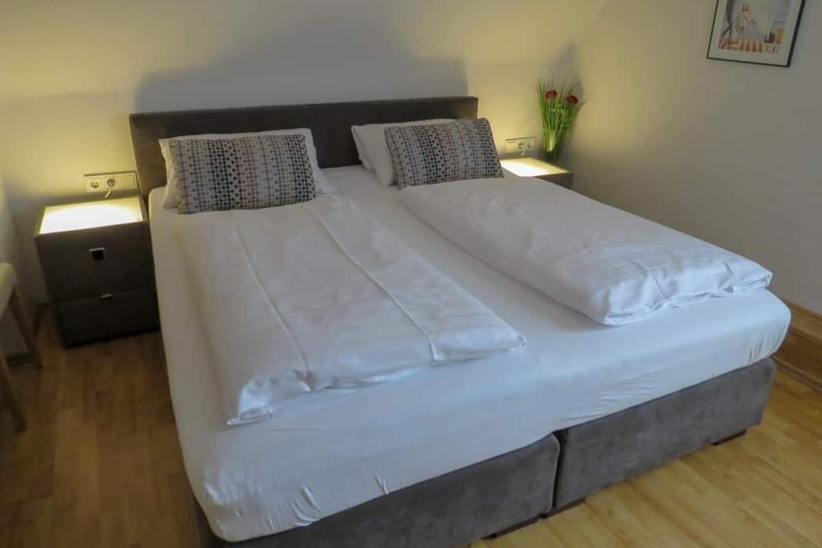Un letto matrimoniale rifatto con cura, lenzuola bianche e due cuscini, un comodino con una lampada e un piccolo vaso con fiori sul ripiano.