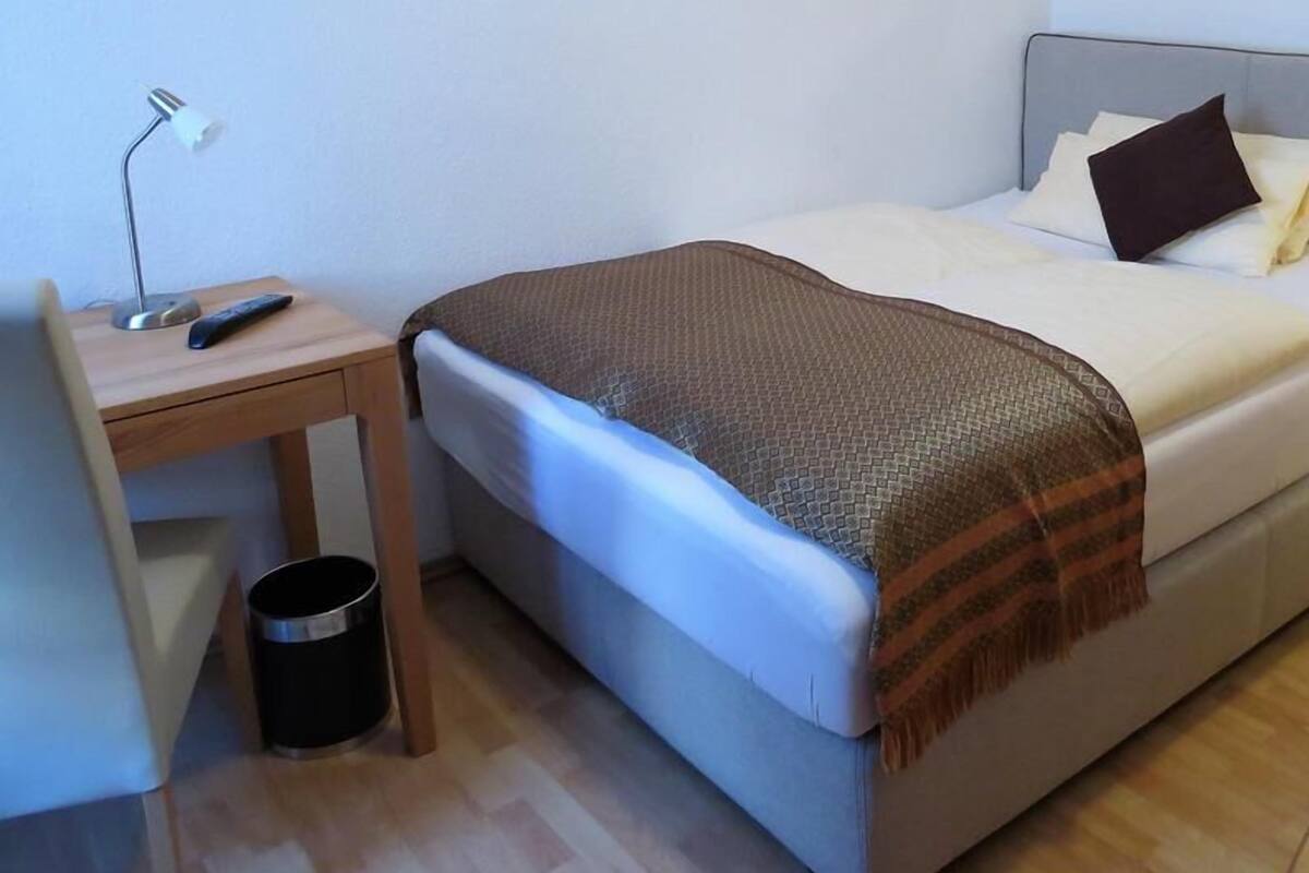 Una camera da letto con un letto, una scrivania, una sedia, una lampada e un cestino.
