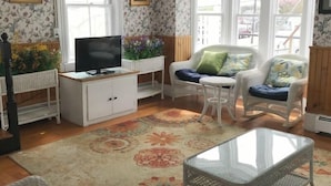A cozy room with floral wallpaper, a television on a white cabinet, a wicker chair with cushions, and a glass coffee table.