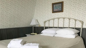 A neatly made bed with white linens and pillows, a stack of folded towels, a bedside lamp, and a floral-patterned rug.