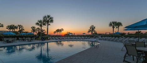 A poolside area with lounge chairs and umbrellas, palm trees, and a sunset view.