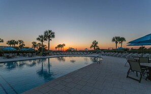 A poolside area with lounge chairs and umbrellas, palm trees, and a sunset view.