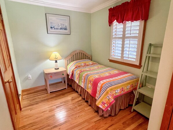 A bedroom with a wooden bed, a nightstand with a lamp, a window with blinds, a shelf, and a framed picture on the wall.