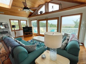 A spacious living room with a large sofa, a coffee table, and a television. There are large windows offering a view of the outdoors.