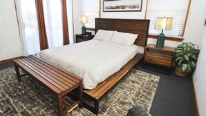 Un dormitorio con una cama grande, un banco de madera, dos mesitas de noche con lámparas, un cuadro en la pared y una planta en maceta.