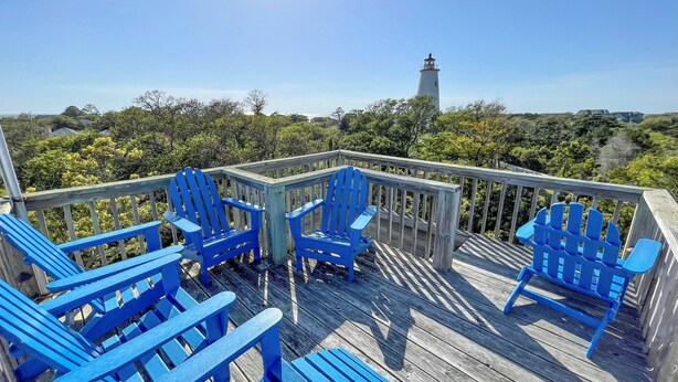 Una terraza de madera con sillones Adirondack azules y un faro al fondo.