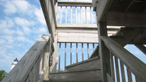 Una escalera de madera que sube a un balcón con vistas a un faro al fondo.