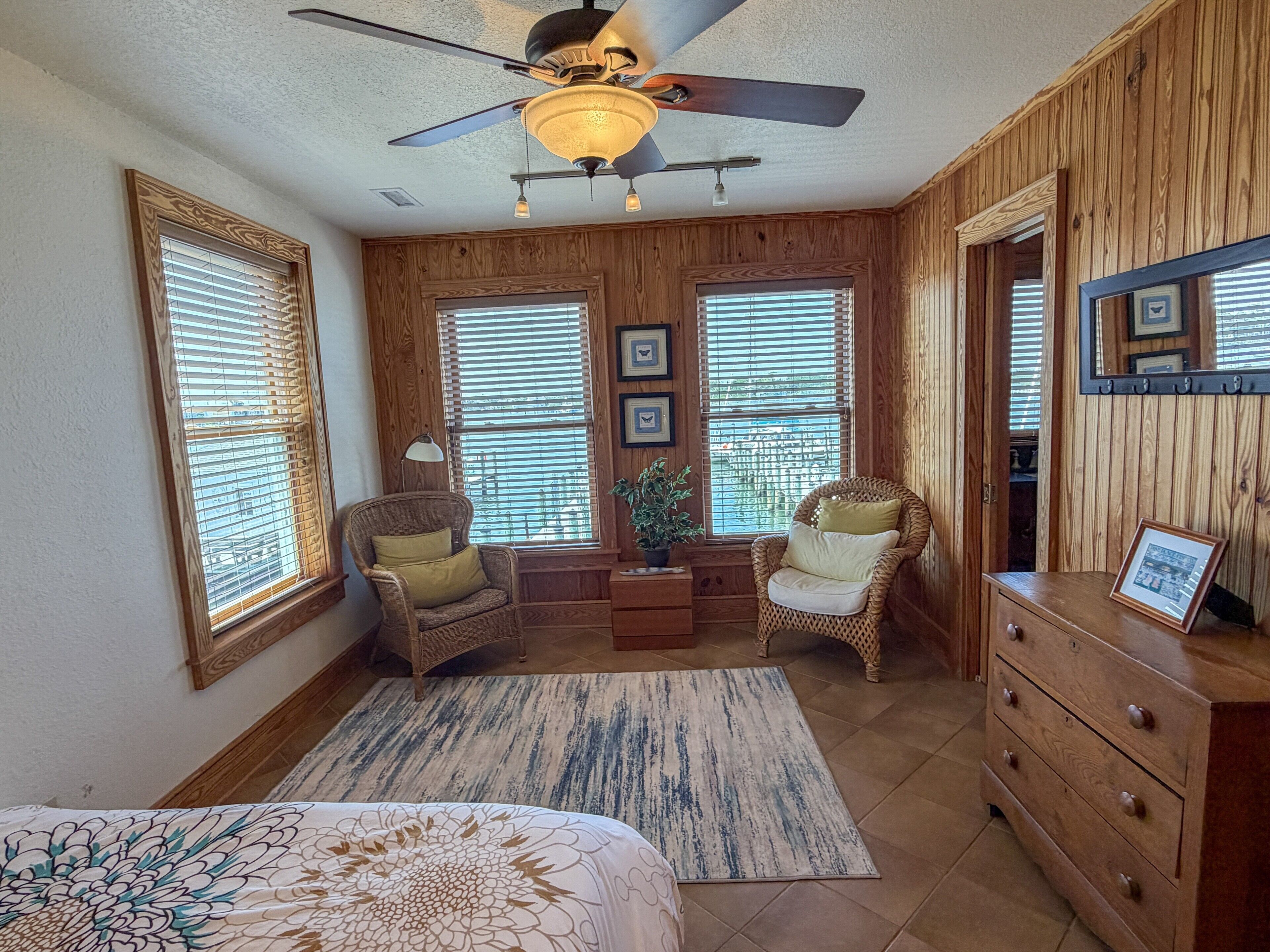 A bedroom with a bed, two wicker chairs, a dresser, and a ceiling fan.