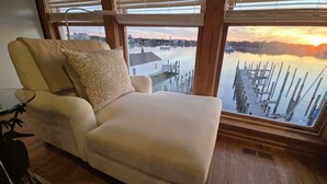 Ein Zimmer mit großem Fenster und Blick auf eine Marina bei Sonnenuntergang, ausgestattet mit einem gepolsterten Sessel und einem kleinen Tisch.