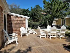 Eine Holzterrasse mit weißen Adirondack-Stühlen und einem Tisch, umgeben von Bäumen und einem Gebäude.