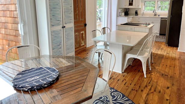 A modern kitchen with white cabinets, a central island, and a dining area with a wooden table and chairs. - Bragg-Tolson House (Formerly Serendipity): Historic Home with Modern Convenience (Ocracoke)