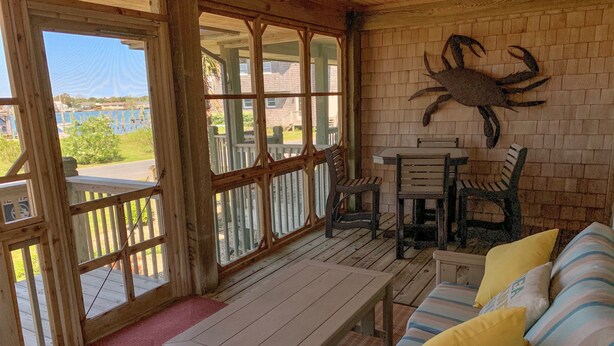 A covered porch with a wooden table and chairs, a cushioned bench with pillows, and a large crab decoration on the wall.