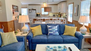 A living room with a blue sofa, yellow cushions, a white coffee table with decorative items, and a kitchen in the background.