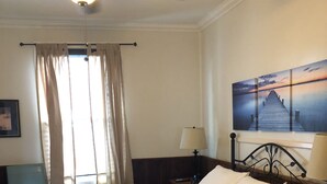 Une chambre d’hôtel avec un grand lit, un ventilateur de plafond, une table de chevet, une lampe, une chaise et un tableau au mur.