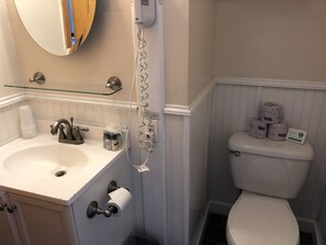 A bathroom with a white sink, a glass shelf, a toilet, and a paper towel dispenser. - Water Street Inn (Block Island)