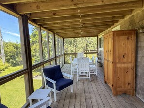 A covered porch with wooden flooring, white furniture, and large windows offering a view of greenery. - Olivella: Pet friendly, artistic, eclectic artwork, decks, porches. (Ocracoke)