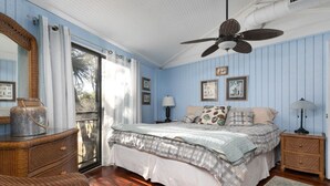 A bedroom with a large bed, two bedside tables with lamps, a ceiling fan, a wicker chair, a large mirror, and a sliding glass door leading to a balcony.