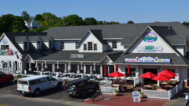 Un bâtiment abritant une boulangerie et un magasin de glaces, avec une terrasse extérieure dotée de parasols et une clôture blanche en bois.