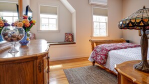 Une chambre avec un lit en bois, une table de chevet avec une lampe, une commode ornée d’un vase de fleurs et une fenêtre avec des stores.