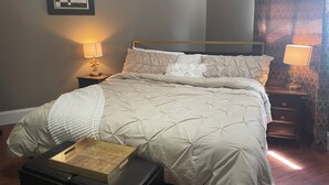 Un lit bien fait, avec une tête de lit matelassée, deux lampes de chevet, un tableau encadré au mur et une petite table sur laquelle se trouvent un livre et une boîte.