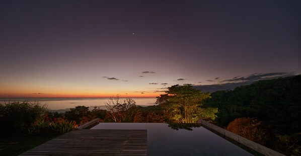 Une terrasse en bois mène à une piscine d’où l’on peut admirer le coucher de soleil sur l’océan et une colline boisée.