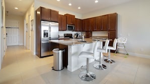 A modern kitchen with a stainless steel refrigerator, a microwave, and a coffee maker. There are three white bar stools and a high chair.