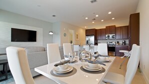 A modern dining area with a white table set for four, surrounded by white chairs. A kitchen with dark wooden cabinets and stainless steel appliances is visible in the background.