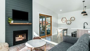 A modern living room with a fireplace, a flat-screen TV, a sofa, a dining area, and a kitchen with a pendant light. - Colorado Modern Cabin (Fort Collins)