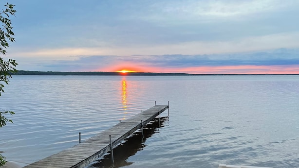 A beach with a wooden dock extending into calm waters, a fire pit, and a boat on the sand. - Sunset Beach - Log Home on Beautiful Indian Lake (Manistique)