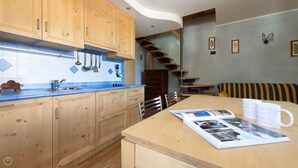 A modern kitchen with wooden cabinets, a blue tiled backsplash, and a wooden dining table with magazines and a coffee mug. - Meralda 2 SCOIATTOLO (Valfurva)