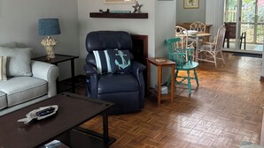 Un salon comprenant un canapé, un fauteuil bleu, une table basse et un coin repas avec des chaises, et donnant sur l’extérieur grâce à une porte vitrée.