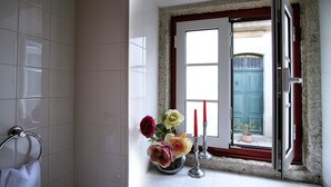 A bathroom with white tiles, a towel rack, a window with a view of a blue door and a potted plant outside. - Casa de David - Apartment in Monsanto (Monsanto, Idanha-a-Nova)