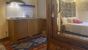 A room with a wooden cabinet, a kitchen area with a stove and kettle, a patterned rug, and a bed with pillows. - Casa de David - Apartment in Monsanto (Monsanto, Idanha-a-Nova)