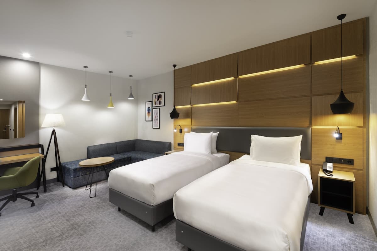 A modern hotel room with two beds, a desk, a sofa, and a small table.