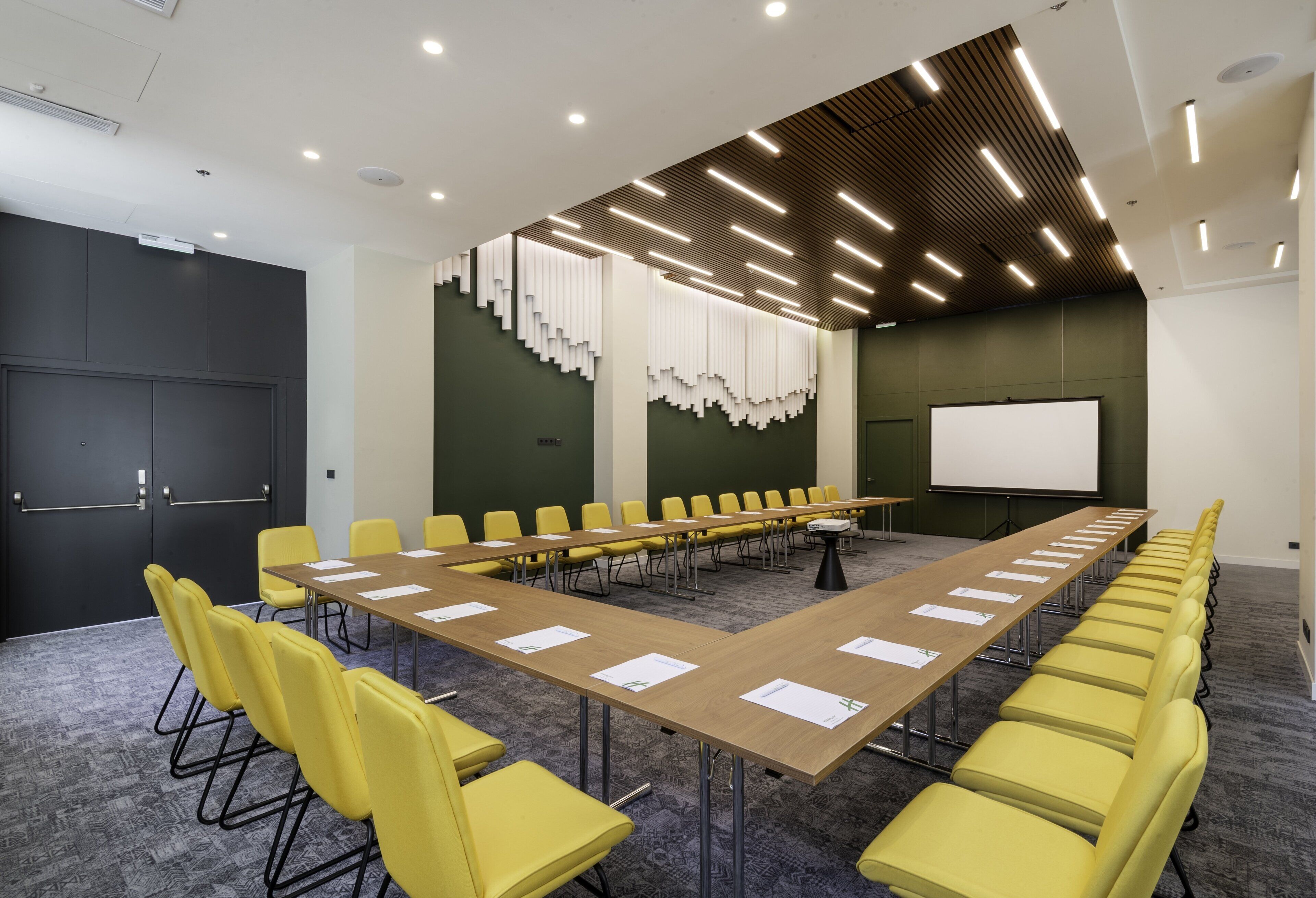 A conference room with a long table, yellow chairs, a projector screen, and a dark-colored door.