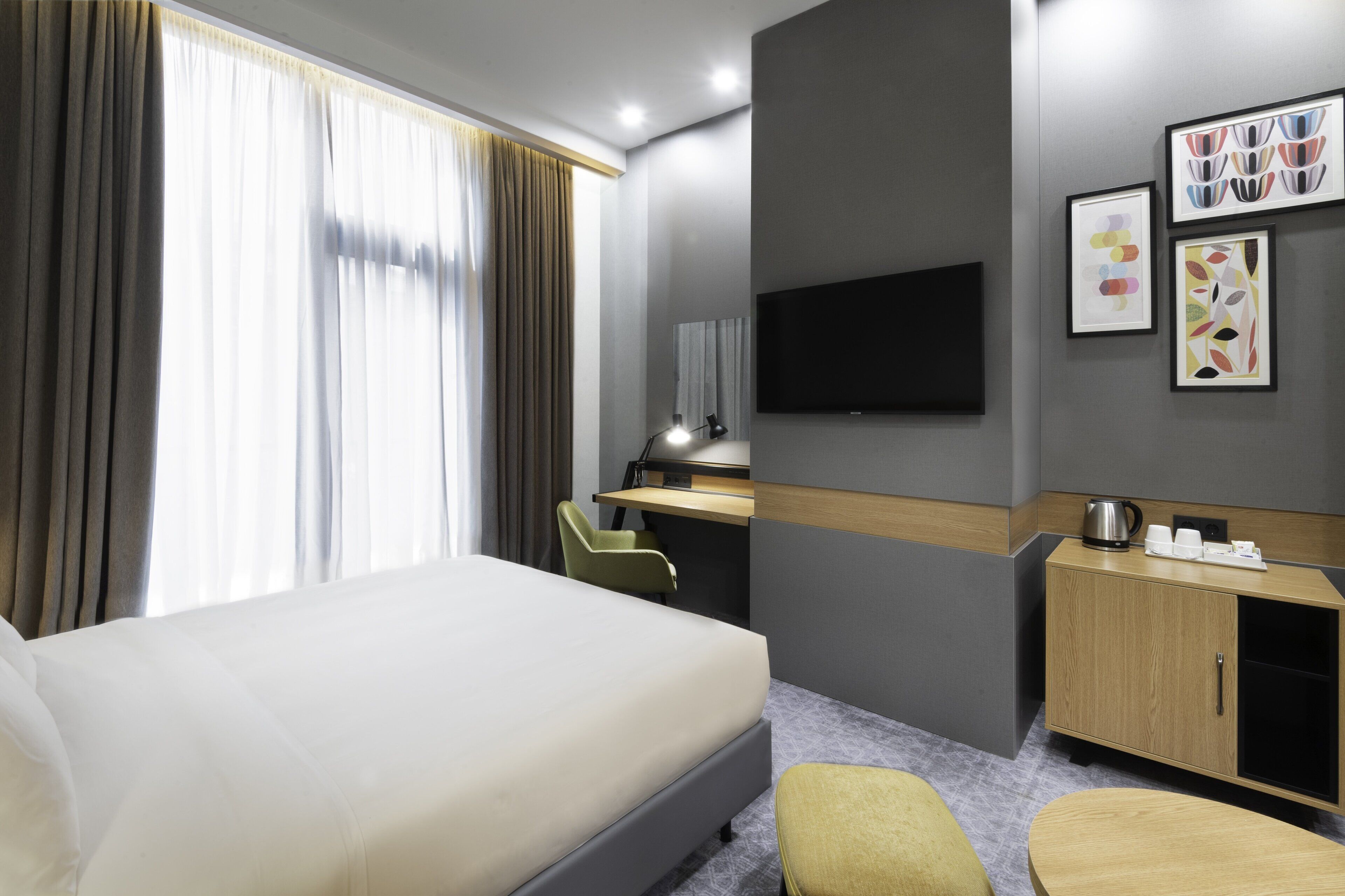 Ein modernes Hotelzimmer mit einem großen Bett, einem Schreibtisch, einem Stuhl, einem an der Wand montierten Fernseher und einer kleinen Kommode mit Wasserkocher und Tassen.