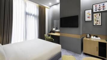 Ein modernes Hotelzimmer mit einem großen Bett, einem Schreibtisch, einem Stuhl, einem an der Wand montierten Fernseher und einer kleinen Kommode mit Wasserkocher und Tassen.