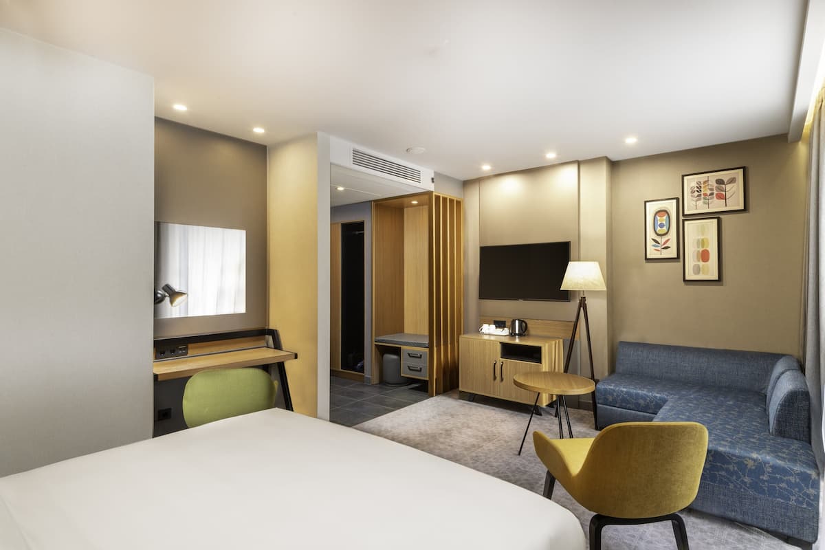 A hotel room with a bed, a desk, a chair, a sofa, a TV, and artwork on the wall.
