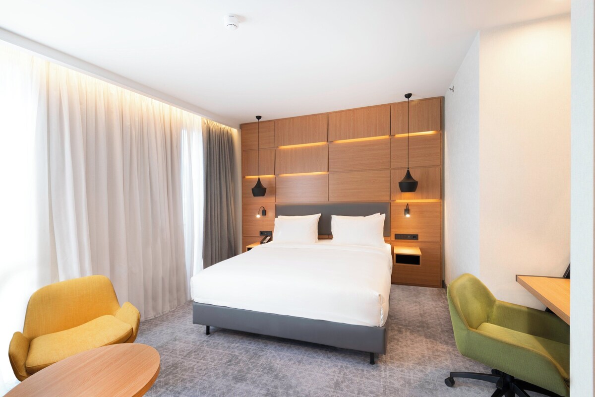 Een moderne hotelkamer met een groot bed, twee stoelen, een bureau en een groot raam met gordijnen.