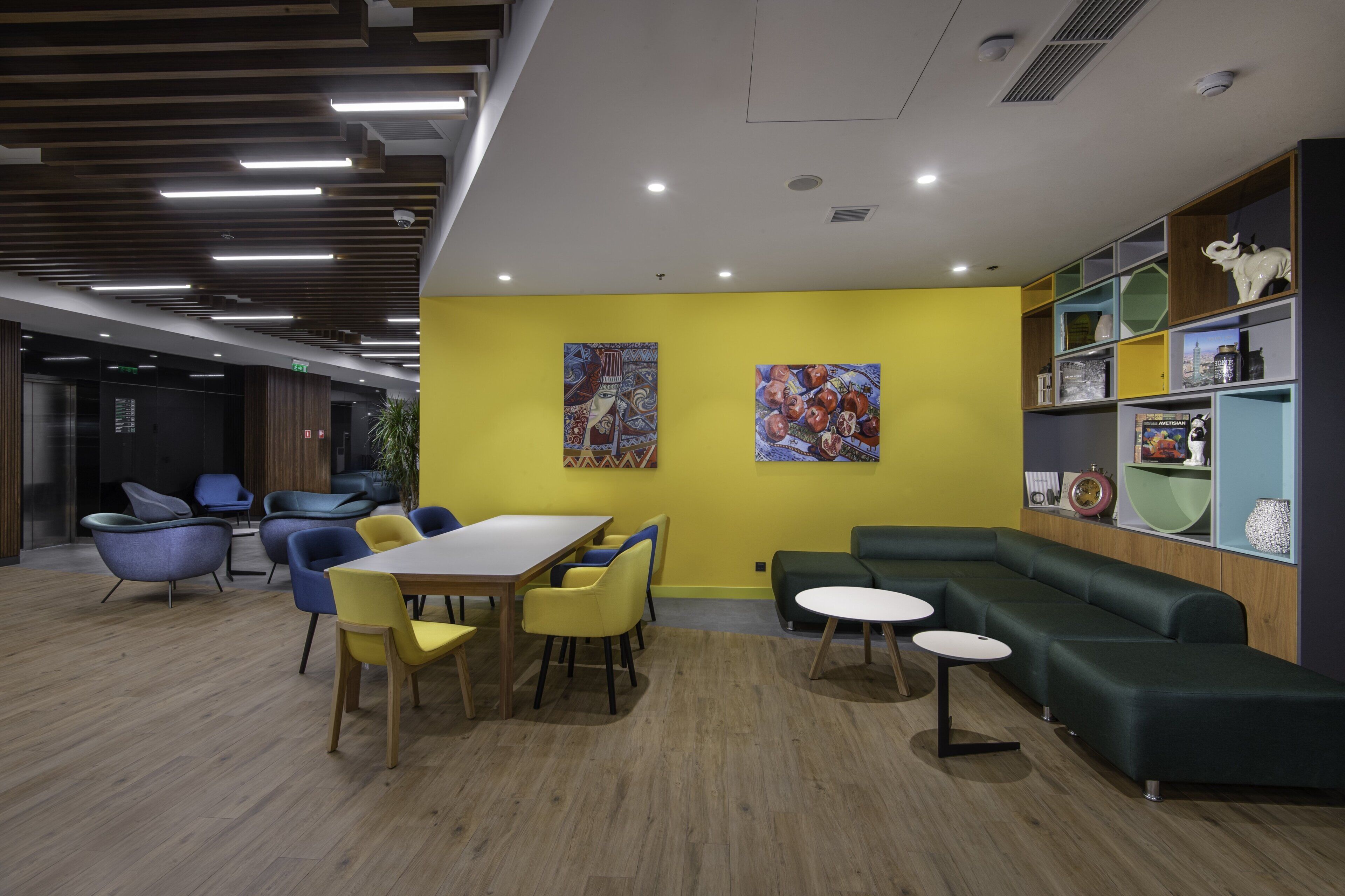 A modern interior with a yellow accent wall, a dining area with yellow chairs, a green sofa, and a bookshelf with decorative items.
