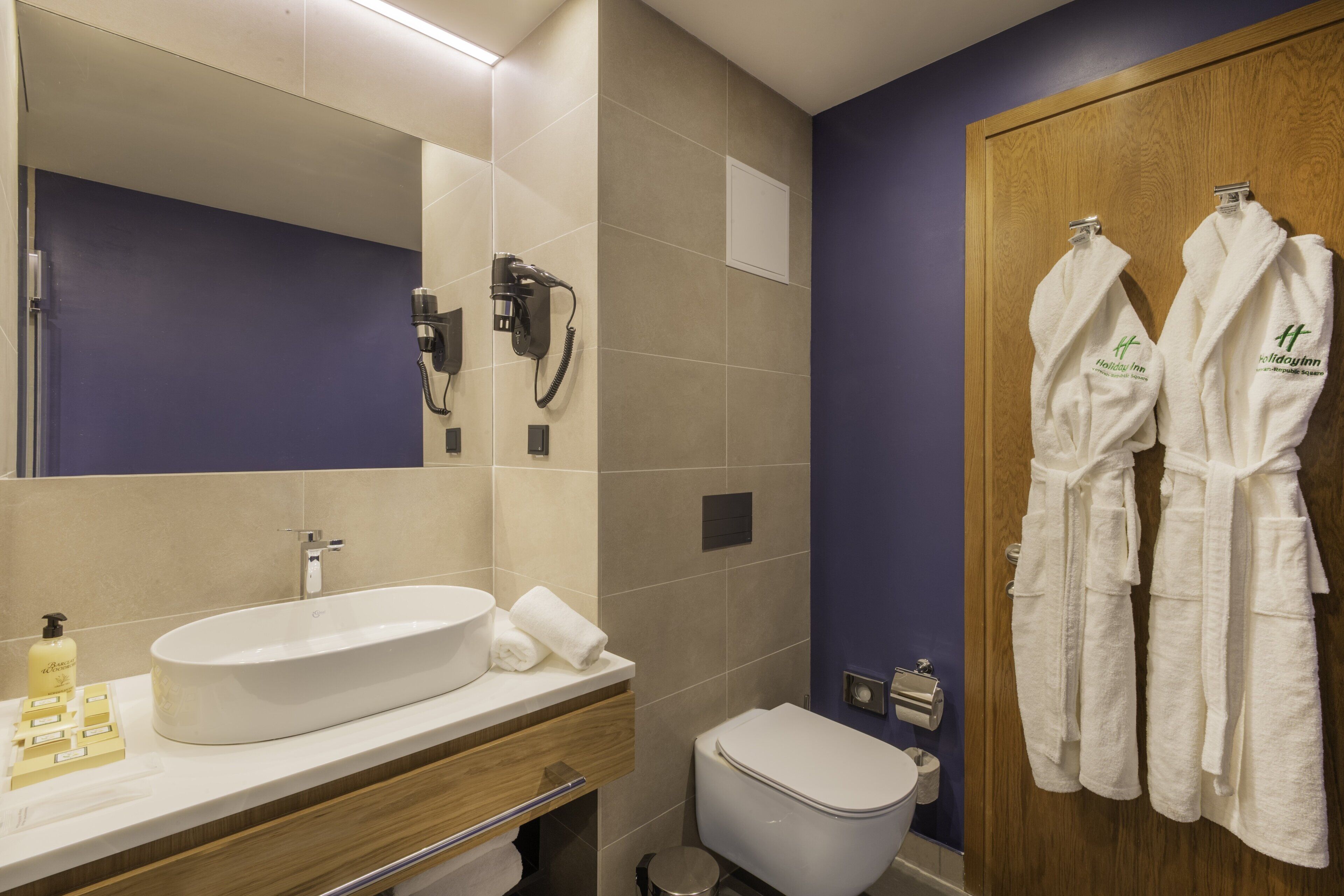 A bathroom with a large mirror, a sink with a bottle of soap, a toilet, and two white robes hanging on a wooden door.