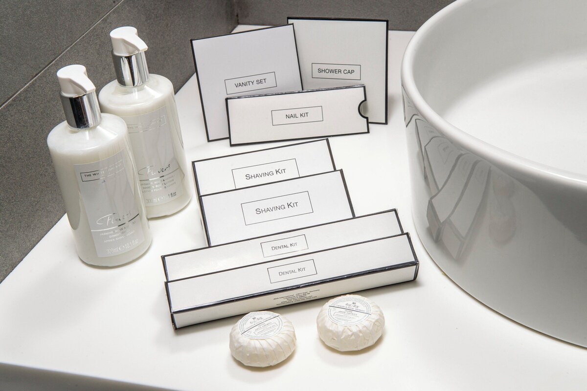 Een badkamermeubel met een groot wastafel, drie flesjes lotion en diverse toiletartikelen sets, gelabeld 'Douchemuts', 'Nagelset', 'Scheerset' en 'Tandheelkundige set'.