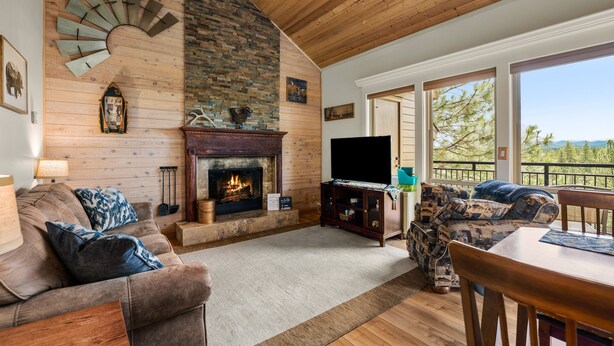 A cozy living room with a fireplace, wooden ceiling, and large windows offering a view of trees. - Dog friendly at the Seventh Mountain Resort! (Bend)