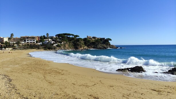 Una playa con costa rocosa, olas rompiendo sobre la arena y edificios al fondo.