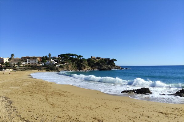 A beach with a rocky shoreline, waves crashing onto the sand, and buildings in the background.