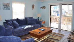 Sala de estar com um sofá azul, uma mesa de centro de madeira e vista para uma casa azul através de uma porta de vidro deslizante.