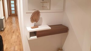Un passadís amb sòl de fusta, paret blanca amb una obra d'art emmarcada, prestatge de fusta amb una fulla decorativa, i un petit vas al terra.