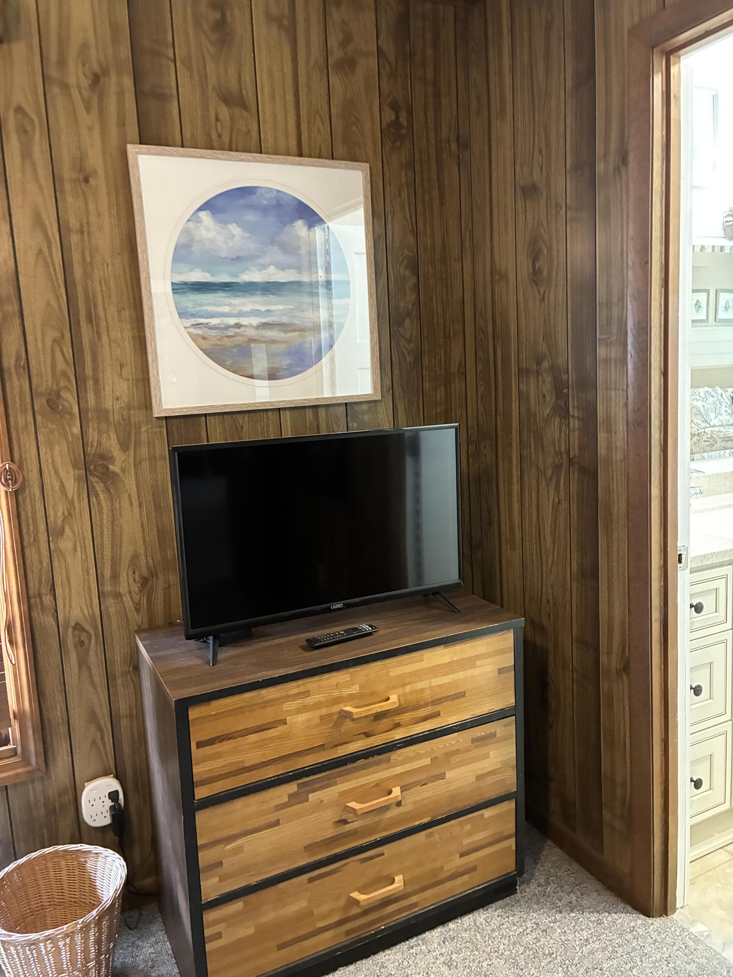 A wooden dresser with a flat-screen TV, a framed picture of a beach, and a wicker basket.