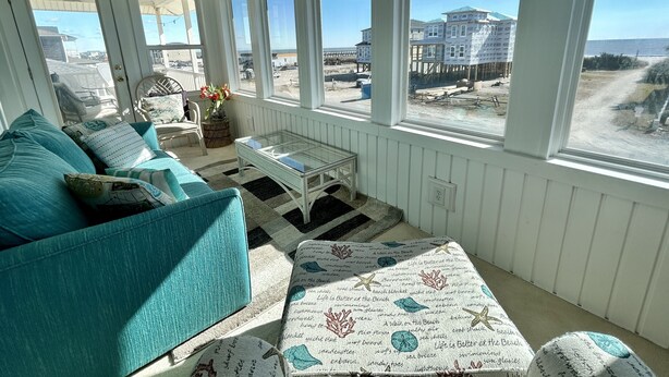 A sunlit room with a view of the beach, featuring a teal sofa, a glass coffee table, and a patterned rug. - Pier View: Cozy Second Row Beach Cottage with Sunroom, Great Views and Easy Beach Access (Brunswick)
