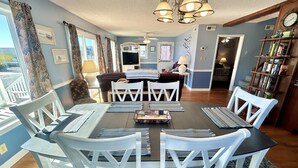 A dining area with a round table, white chairs, and blue placemats. - Pier View: Cozy Second Row Beach Cottage with Sunroom, Great Views and Easy Beach Access (Brunswick)